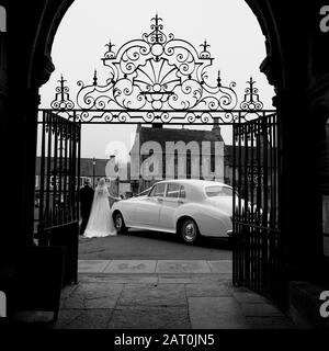 Ein Bride und Ein Bräutigam, die die Kirche verlassen, als sie mit ihrem Chauffeur und dem Auto heiraten, das auf sie draußen wartet Stockfoto