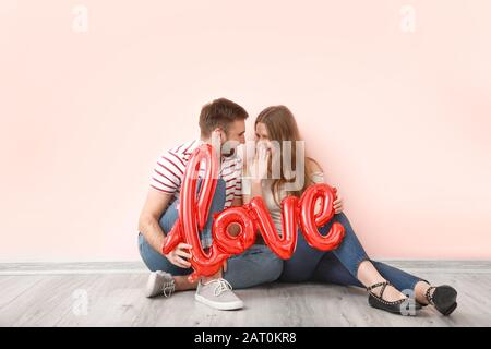 Glückliches junges Paar mit Ballon in Form von WORTLIEBE in der Nähe der Farbwand sitzend. Valentinstag gefeiert Stockfoto