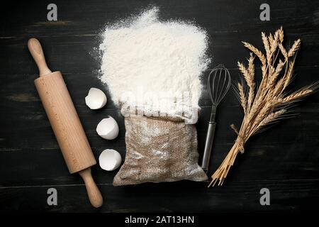Beutel mit Mehl, Utensilien, Eierschale und Weizenpikeletten auf dunklem Hintergrund Stockfoto
