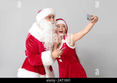 Junges Paar, das sich als Weihnachtsmann verkleidet hat und selfie auf hellem Hintergrund nimmt Stockfoto