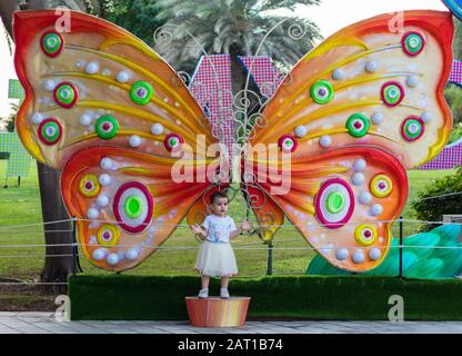 Ein süßes kleines Mädchen in einem weißen Kleid, das vor dem riesigen Schmetterling im Dubai Glow Garden posiert. Stockfoto