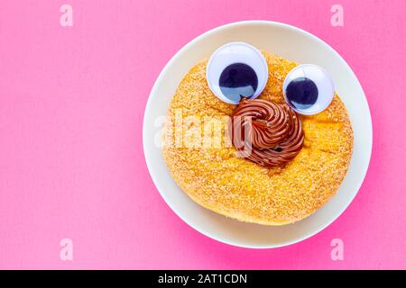 Ein Tim Hortons Dulce de Leche Donut mit Black Wiggle Googly Augballs auf einem Teller mit pinkfarbenem Hintergrund Stockfoto