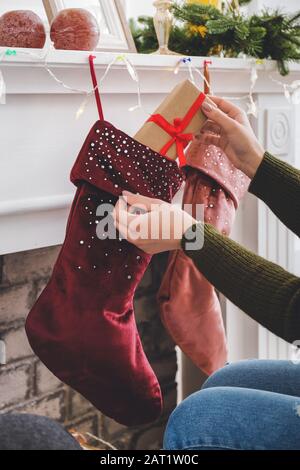 Frau, die Geschenke in Weihnachtssocken legt, die zu Hause am Kamin hängen Stockfoto