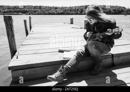 Kaukasisches Teenager-Mädchen in Brille spielt Ukulele auf einem Holzsteg, Schwarz-Weiß-Außenfoto Stockfoto