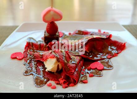 Platte mit Himbeere, roter Kreppe mit Schokoladensauce und pinkfarbenen, herzförmigen Süßigkeiten Stockfoto