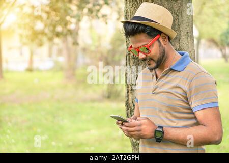 Fröhlicher inder, der sein Handy sieht und lächelt - Junger asiate, der online im Park im Freien textet - Neues Technologietrends Suchtkonzept - Krieg Stockfoto