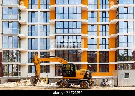 Ein gelber Bagger, der vor einem Wohnhochhaus mit Glasfenstern arbeitet. Stockfoto