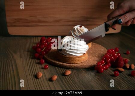 Zwei Cupcakes mit frischen Beeren und Mandeln auf einem Korkständer und menschliche Hand mit einem großen Messer Stockfoto