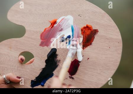 Nahaufnahme der jungen Künstlerin, die an der Malerei im Freien arbeitet. Sie hält Ölfarben, Künstlerbürsten, Leinwand und Palette. Sie mischt Farben ein Stockfoto