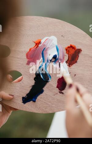 Nahaufnahme der jungen Künstlerin, die an der Malerei im Freien arbeitet. Sie hält Ölfarben, Künstlerbürsten, Leinwand und Palette. Sie mischt Farben ein Stockfoto