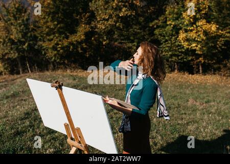 Oung weibliche Künstlerin, die im Freien, in der Herbstlandschaft, Malerei beschäftigt. Sie steht vor der Leinwand und Zeichnung.Sie hält Ölfarben, Künstlerbürste Stockfoto