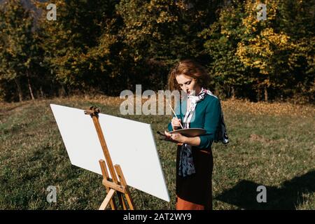 Oung weibliche Künstlerin, die im Freien, in der Herbstlandschaft, Malerei beschäftigt. Sie steht vor der Leinwand und Zeichnung.Sie hält Ölfarben, Künstlerbürste Stockfoto