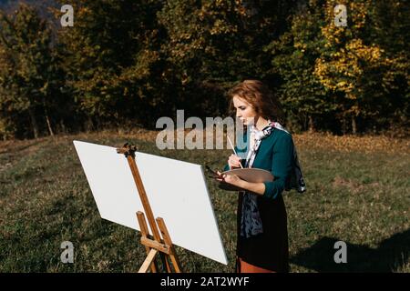 Oung weibliche Künstlerin, die im Freien, in der Herbstlandschaft, Malerei beschäftigt. Sie steht vor der Leinwand und Zeichnung.Sie hält Ölfarben, Künstlerbürste Stockfoto