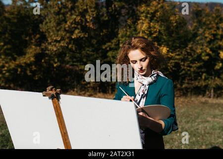 Oung weibliche Künstlerin, die im Freien, in der Herbstlandschaft, Malerei beschäftigt. Sie steht vor der Leinwand und Zeichnung.Sie hält Ölfarben, Künstlerbürste Stockfoto