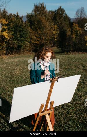 Oung weibliche Künstlerin, die im Freien, in der Herbstlandschaft, Malerei beschäftigt. Sie steht vor der Leinwand und Zeichnung.Sie hält Ölfarben, Künstlerbürste Stockfoto