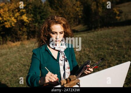 Oung weibliche Künstlerin, die im Freien, in der Herbstlandschaft, Malerei beschäftigt. Sie steht vor der Leinwand und Zeichnung.Sie hält Ölfarben, Künstlerbürste Stockfoto