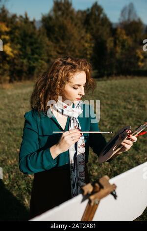 Oung weibliche Künstlerin, die im Freien, in der Herbstlandschaft, Malerei beschäftigt. Sie steht vor der Leinwand und Zeichnung.Sie hält Ölfarben, Künstlerbürste Stockfoto