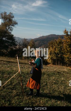 Oung weibliche Künstlerin, die im Freien, in der Herbstlandschaft, Malerei beschäftigt. Sie steht vor der Leinwand und Zeichnung.Sie hält Ölfarben, Künstlerbürste Stockfoto