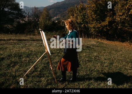 Oung weibliche Künstlerin, die im Freien, in der Herbstlandschaft, Malerei beschäftigt. Sie steht vor der Leinwand und Zeichnung.Sie hält Ölfarben, Künstlerbürste Stockfoto