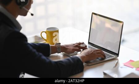 Seitenschuss des jungen, intelligenten Bedieners mit Headsets, die in einem Callcenter arbeiten, während er auf dem weißen Laptop mit leerem Bildschirm tippt und am Schreibtisch sitzt. Stockfoto