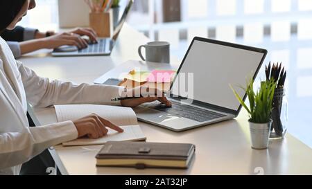 Zugeschnittene junge Geschäftsfrau, die auf dem weißen Laptop mit leerem Bildschirm tippt, indem sie den Informationen aus den Notizen folgt und am Schreibtisch sitzt Stockfoto