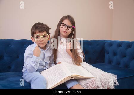 Bruder und Schwester sitzen auf dem Sofa in einer Papierbrille und lesen spielerisch ein Buch Stockfoto