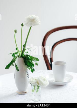 Eleganter Tisch und ein Sessel mit weißen Ranunculus-Blumen und einer Tasse Kaffee. Guter Vormittagstisch für den Valentinstag oder Den Tag Der Frau Stockfoto