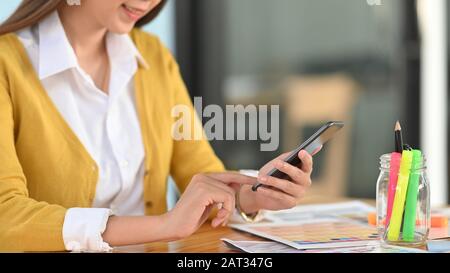 Zugeschnittene Aufnahme der jungen Designerin, die Smartphone in der Hand hält und mit dem Holztisch als Hintergrund darauf zeigt. Stockfoto