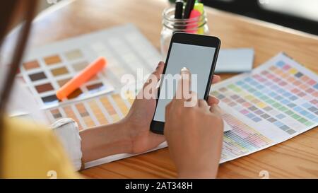 Beschnittener Schuss der jungen Designerin, die weißes leeres Bildschirm-Smartphone in der Hand hält und mit dem Finger darauf zeigt, mit dem Holztisch als Hintergrund Stockfoto