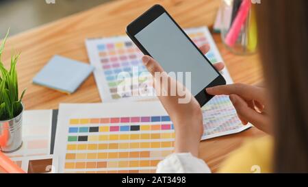 Beschnittener Schuss der jungen Designerin, die weißes leeres Bildschirm-Smartphone in der Hand hält und mit dem Finger darauf zeigt, mit dem Holztisch als Hintergrund. Stockfoto