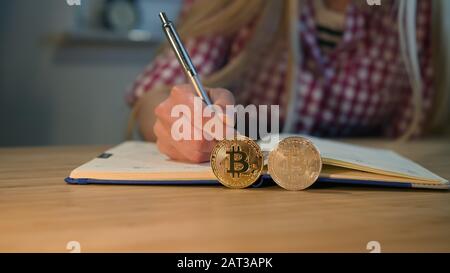 Schnittansicht weiblicher Hände in kariertem Hemd mit glänzenden Metallstiften und Schreiben in den Tagesplaner mit Bitcoins, die nachts an Kanten im Vordergrund platziert werden. Stockfoto