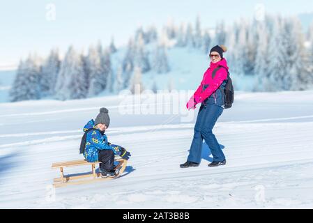 Mutter schleppt mit Junge den Schneeschlitten. Berg im Hintergrund. Das Konzept hält Kinder im Winter gesund Stockfoto