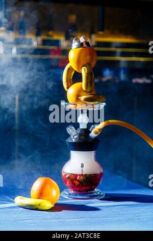 Hookah mit einem runden transparenten Kolben, in dem eine rote Flüssigkeit vorhanden ist. Kapuzenschüssel mit Orangen. Viel Rauch. Schwarzer Hintergrund. Gelbes Mundstück. Stockfoto