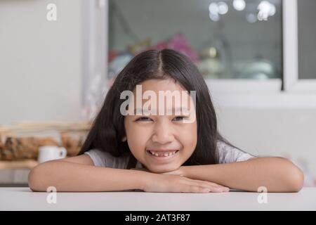 Das kleine Mädchen legte ihr Kinn auf beide Hände und lächelte, um ihre gebrochenen Zähne auf dem weißen Kochtisch in der Küche zu sehen. Stockfoto