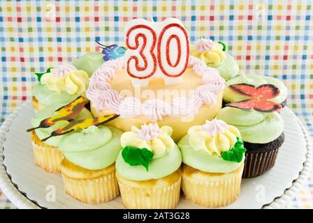 Blick auf einen Geburtstagskuchen und Cupcakes in pastellfarbenem Buttercremeficing auf einem Kuchenständer. Wachskerze Nummer 30 steht oben. Stockfoto