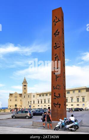 Trani, Apulien/Italien - 06.08.24: Moderne Obelisk-Dekoration am Piazza Re Manfredi mit der Kathedrale St. Nikolaus Der Pilgrim im Hintergrund entlang der adriatischen Seepromenade in der Altstadt von Trani. Stockfoto