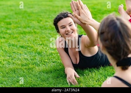 Zwei glückliche Frauen in Sportbekleidung, die nach Übungen auf dem Gras liegen, klatschen die Hände, draußen. - Bild Stockfoto