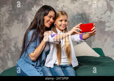 Fröhliche Mutter zart umarmt ihre hübsche Tochter. selfie auf dem Smartphone machen, auf dem Bett sitzen. Abbildung: Konzept für Elternschaft Stockfoto