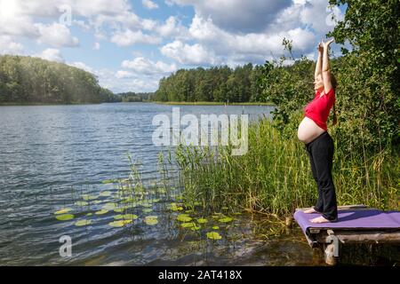 Junge schwangere Frau schleift an einem Holzsteg an einem Waldsee während des warmen Sonntags, der Fitnessübungen macht - gesunder Schwangerschafts-Lebensstil c Stockfoto