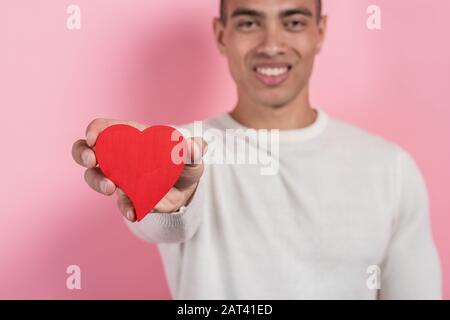 Mulatto-Mann hält ein Herzobjekt in der geraden Hand. Fokus auf Herz, verschwommener Hintergrund.- Bild Stockfoto