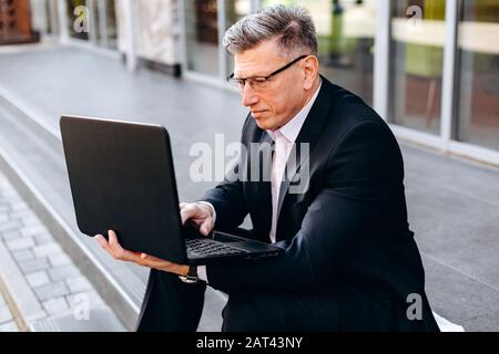 Porträt des älteren Mannes im Anzug auf dem Straßenbelag sitzend und einen Laptop halten und draußen tippen. - Bild Stockfoto