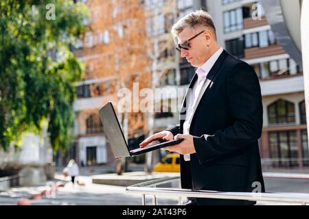 Ein seriöser, leitender Geschäftsmann in Sonnenbrillen, der einen Laptop hält und in ihm im Freien arbeitet. - Bild Stockfoto