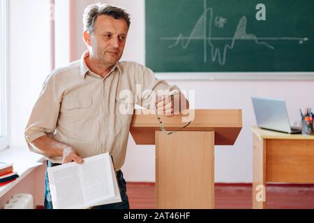 Lächelnder Lehrer mittleren Alters, der ein Buch stehend hält und sich zum Dreigestirn neigt. Konzeptbildung Stockfoto