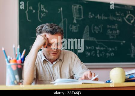 Mittelalterslehrer in Brille liest Lehrbuch aufmerksam im Klassenzimmer und lächelt.- Bild Stockfoto