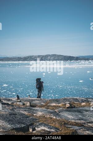 Junger weißer kaukasier mit Rucksack auf einer Wandertour in Grönland mit Eisbergen und Blick auf den Atlantik. Arktische Landschaft im Sommer. In Der Nähe Von Arcti Stockfoto