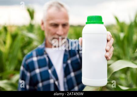 Bauer mittleren Alters auf einem Feld, das eine Flasche mit chemischen Düngern zeigt. Konzentrieren Sie sich auf die Flasche Stockfoto