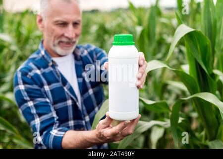 Bauer mittleren Alters steht auf einem Feld, der auf eine Flasche mit chemischen Düngern in seinen Händen blickt. Düngerflasche verspottern Stockfoto