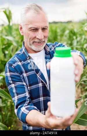 Bauer mittleren Alters steht auf einem Feld, der auf eine Flasche mit chemischen Düngern in seinen Händen blickt. Düngerflasche verspottern Stockfoto