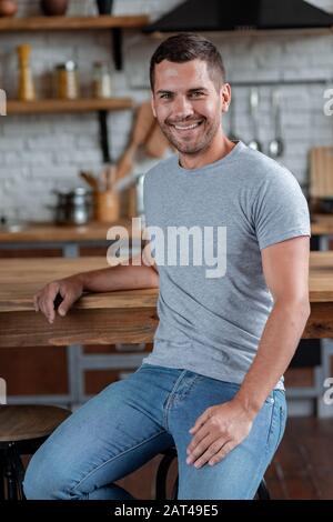 Ansehnlicher Mann sitzt auf dem Stuhl, der sich auf den Tisch gelehnt hat und lächelt mit Blick auf die Kamera.- Bild Stockfoto