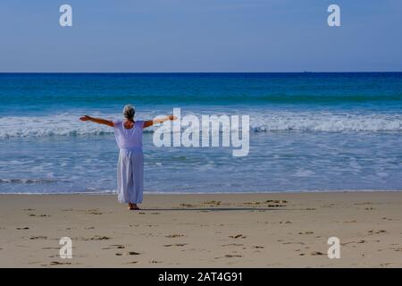 Ältere Frau mit grauem Haar, die Qi Gong und Yoga am Strand von Tarifa macht Stockfoto